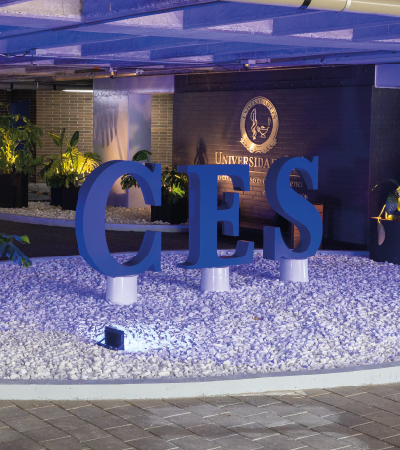 Zona exterior iluminada con un gran letrero azul que forma las letras ‘CES’, ubicado sobre un área cubierta de piedras blancas y frente a un muro con el nombre y escudo de la universidad.