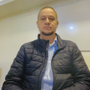 La imagen muestra a una persona sentada en un espacio interior, vistiendo una chaqueta acolchada negra sobre una camisa azul clara. El fondo es una pared clara con iluminación suave.