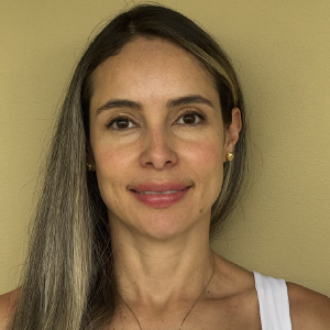 La imagen muestra a una persona de pie frente a una pared de color beige, con el cabello largo y lacio peinado hacia un lado. Lleva una blusa sin mangas de color blanco y unos aretes dorados pequeños.