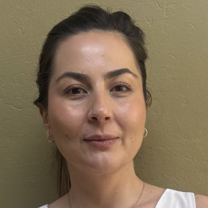 La imagen muestra a una persona de pie frente a una pared beige, con el cabello recogido hacia atrás. Lleva una blusa blanca sin mangas y unos aretes pequeños dorados.