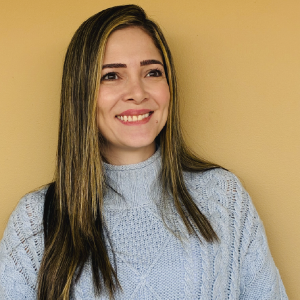La imagen muestra a una persona de pie frente a una pared de color beige, vistiendo un suéter tejido de color gris claro con diseño de trenzas. Tiene el cabello largo, lacio y con mechas claras, peinado hacia adelante sobre los hombros.