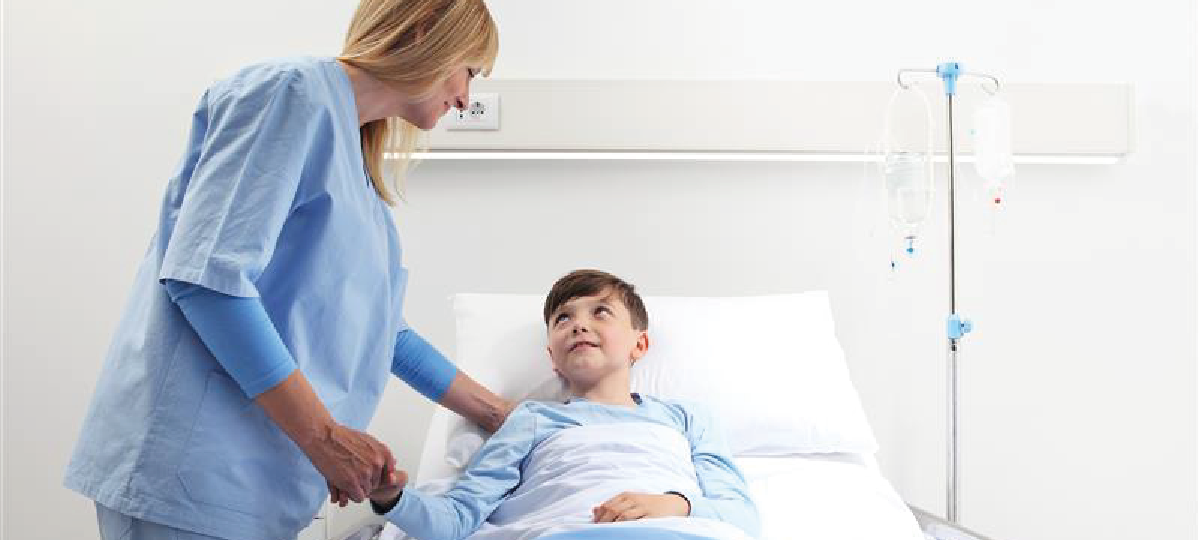 La imagen muestra a una profesional de la salud junto a un paciente recostado en una cama hospitalaria. La profesional sostiene la mano del paciente y se inclina ligeramente hacia él. Ambos visten ropa clínica de color azul claro, y junto a la cama se observa un soporte con bolsas de suero. El entorno es una habitación hospitalaria iluminada y de apariencia limpia.