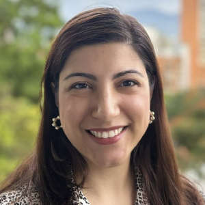 La imagen muestra a una persona de pie en un entorno exterior, con el cabello largo y castaño peinado recto. Viste una blusa con estampado en tonos claros y lleva aretes dorados con detalles en forma de perlas. Al fondo se observan árboles y edificios ligeramente desenfocados.