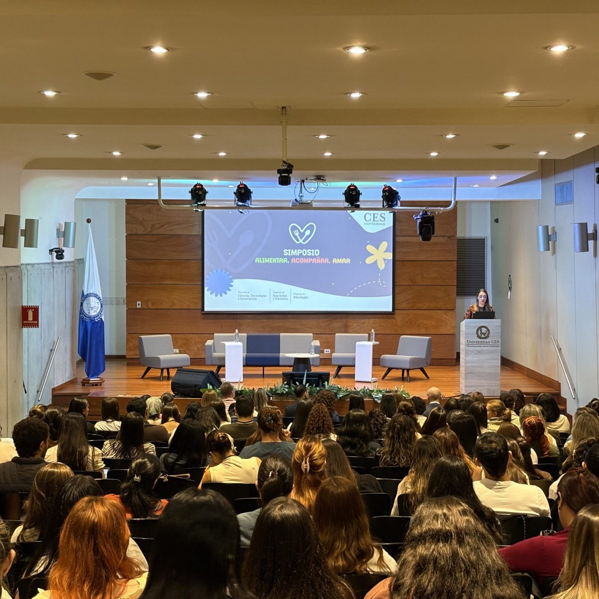 Auditorio lleno de asistentes escuchando una presentación en un escenario con pantalla proyectada y varios asientos dispuestos para un panel. Un expositor habla desde un podio ubicado al lado derecho del escenario.