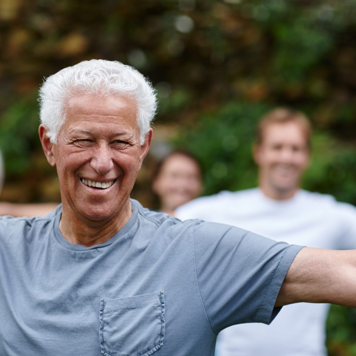 Personas adultas realizando una postura de ejercicio con los brazos extendidos horizontalmente, al aire libre, en un entorno natural con vegetación de fondo.