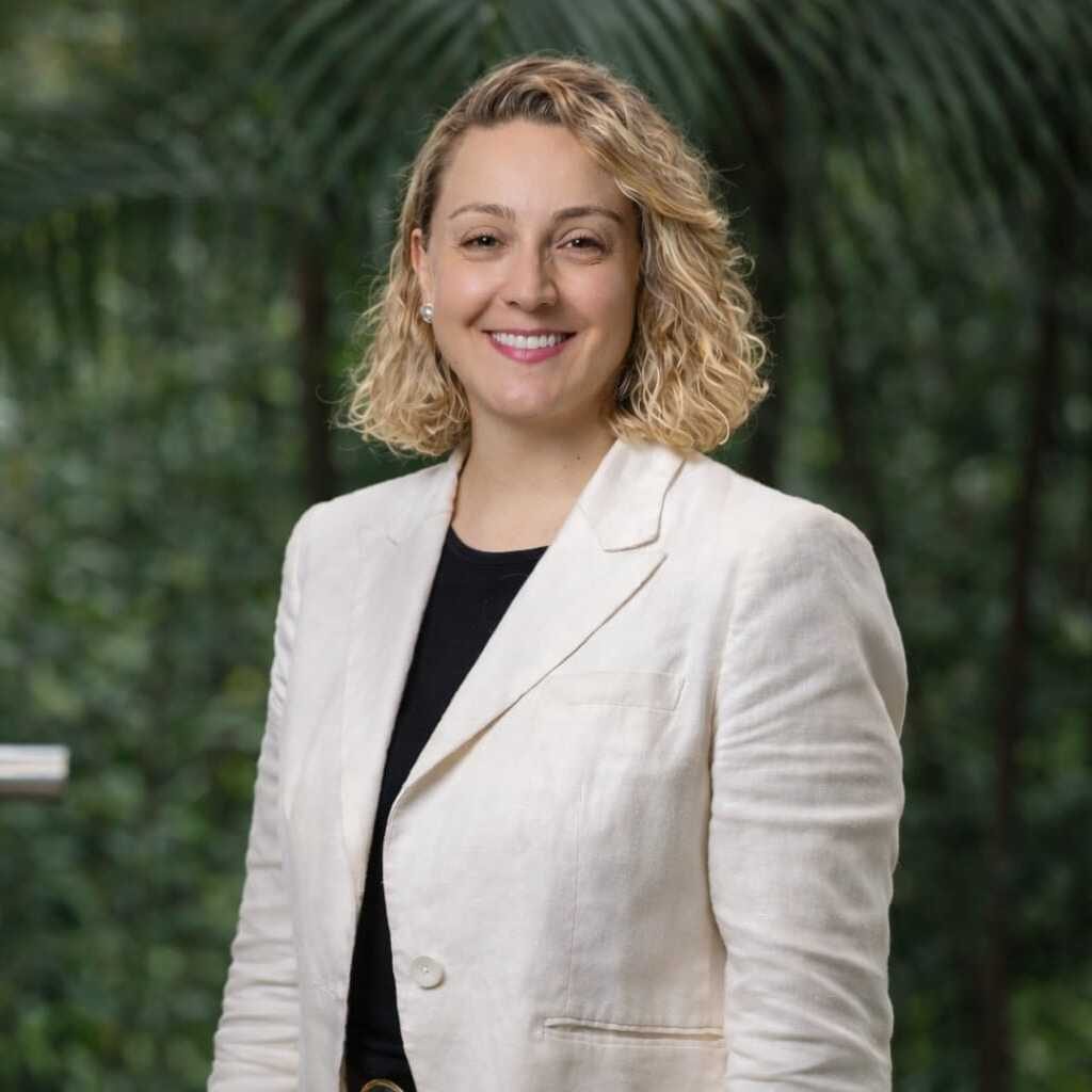 Persona posando de pie, con chaqueta clara y prenda oscura debajo, en un entorno exterior con abundante vegetación y follaje verde desenfocado al fondo.