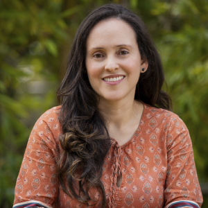 La imagen muestra a una persona sentada en un entorno exterior, vestida con una blusa de color terracota con un patrón decorativo claro. El cabello es largo y oscuro, con ondas suaves hacia un lado. El fondo está compuesto por vegetación verde desenfocada.