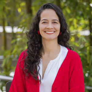 La imagen muestra a una persona sentada en un entorno exterior con vegetación verde desenfocada al fondo. La persona lleva una blusa blanca con cuello en “V” y un cárdigan rojo de textura tejida. El cabello es oscuro, largo y rizado, cayendo sobre los hombros.