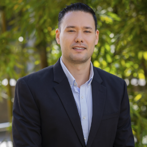 La imagen muestra a una persona de pie en un entorno exterior con vegetación verde desenfocada al fondo. La persona viste una chaqueta formal oscura sobre una camisa clara abotonada. El cabello está peinado hacia atrás y la iluminación es natural.