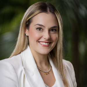 La imagen muestra a una persona en un entorno exterior con vegetación verde desenfocada al fondo. La persona viste un blazer blanco y una blusa clara, y lleva un collar de perlas junto con otro collar dorado con dijes. El cabello es liso, largo y de tono claro.
