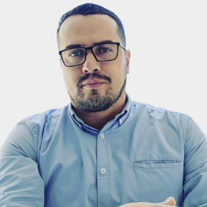 La imagen muestra a una persona sentada, vistiendo una camisa de color azul claro con botones al frente y un bolsillo en el lado izquierdo del pecho. El fondo es completamente blanco, lo que da un aspecto limpio y minimalista. La postura sugiere que los brazos están apoyados o entrelazados frente al cuerpo.