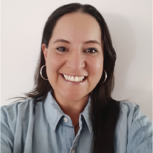 La imagen muestra a una persona frente a una pared blanca, vistiendo una camisa de mezclilla clara abotonada. El cabello es largo y oscuro, con un lado hacia adelante sobre el hombro. La persona lleva aretes grandes circulares de color metálico.