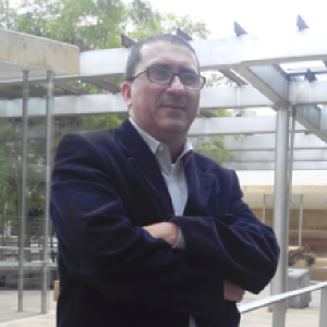 La imagen muestra a una persona de pie en un espacio exterior con estructuras metálicas y techos transparentes. La persona viste un saco oscuro y una camisa clara, y mantiene los brazos cruzados. Al fondo se observan elementos arquitectónicos, barandas y árboles.