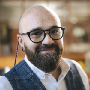 La imagen muestra a una persona en un entorno interior iluminado, vistiendo una camisa blanca y un chaleco de tela a cuadros en tonos azules. También lleva gafas con soporte lateral adornado con un cordón de colores. El fondo presenta estantes y elementos desenfocados propios de un espacio tipo oficina, biblioteca o taller.