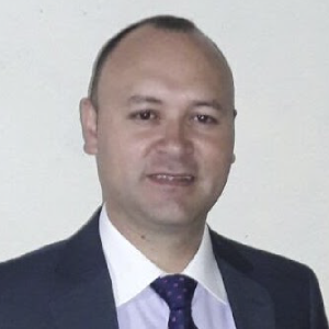 La imagen muestra a una persona frente a un fondo blanco, vistiendo un traje formal compuesto por una chaqueta oscura, una camisa clara y una corbata morada con puntos. El encuadre es de medio cuerpo y la iluminación es uniforme.