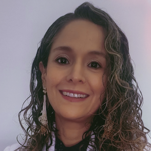 La imagen muestra a una persona frente a un fondo liso de color claro. La persona lleva el cabello largo y rizado, y utiliza aretes largos con detalles colgantes en tonos dorados. También viste una prenda superior clara.