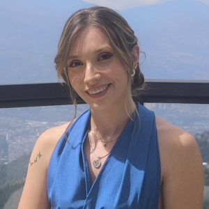 La imagen muestra a una persona en un entorno exterior elevado, con una vista amplia de una ciudad y montañas al fondo. La persona viste una blusa sin mangas de color azul y lleva varios collares dorados. En uno de los brazos se observa un pequeño tatuaje de aves en silueta. El cabello está recogido hacia atrás.