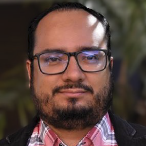 La imagen muestra a una persona en un entorno exterior con vegetación desenfocada al fondo. La persona viste una chaqueta oscura y una camisa de cuadros en tonos rosados y blancos. El encuadre es de medio cuerpo y la iluminación es natural.