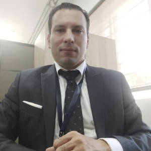 La imagen muestra a una persona en un entorno interior tipo oficina, vestida con un traje formal oscuro, camisa clara y corbata estampada en tonos oscuros. Lleva un cordón portaidentificación colgado sobre el cuello y un pañuelo blanco en el bolsillo del saco. Al fondo se observan divisiones de oficina, muebles de madera y una ventana con persiana que deja entrar luz natural.