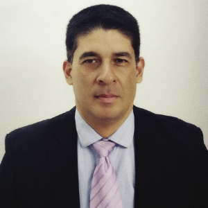 La imagen muestra a una persona frente a un fondo liso de color claro, usando un traje formal oscuro con camisa clara y una corbata de color rosa pálido. El encuadre es de medio cuerpo y la iluminación es uniforme.