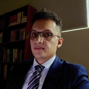 La imagen muestra a una persona en un entorno interior con estanterías de libros en el fondo. La persona viste una camisa clara con una corbata de rayas diagonales en tonos oscuros y un saco formal azul. A un costado entra luz natural por una ventana, creando contraste en la escena.