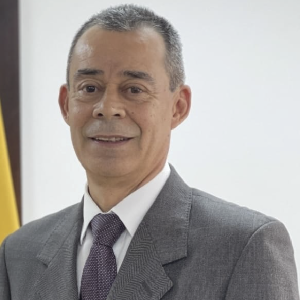 La imagen muestra a una persona vistiendo un traje formal gris con camisa blanca y corbata en tono púrpura oscuro. El fondo es una pared clara y, a un lado, se observa parcialmente una bandera de color amarillo.