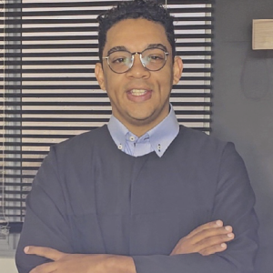 La imagen muestra a una persona de pie en un espacio interior, con los brazos cruzados. La persona viste una camisa clara y una prenda oscura encima, similar a una bata o suéter. El fondo incluye una ventana con persianas horizontales y una pared oscura.
