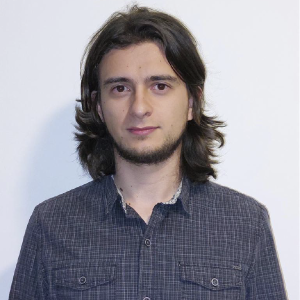 La imagen muestra a una persona de pie frente a un fondo blanco, vistiendo una camisa de cuadros en tonos oscuros. La persona tiene el cabello largo, ondulado y a la altura de los hombros. El encuadre es de medio cuerpo y la iluminación es uniforme.