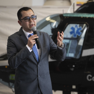 La imagen muestra a una persona usando un traje formal gris y una corbata azul con patrón. La persona sostiene un micrófono mientras realiza un gesto con la otra mano. Al fondo se observa un helicóptero negro con un símbolo médico en color azul sobre una de las ventanas. El entorno sugiere un espacio relacionado con actividades aéromédicas o de transporte especializado.