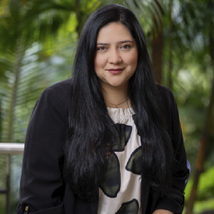 La imagen muestra a una persona en un entorno exterior con vegetación frondosa al fondo. La persona lleva puesta una chaqueta oscura sobre una blusa clara con un diseño grande en tonos oscuros. El cabello es largo, lacio y de color oscuro, cayendo hacia ambos lados sobre los hombros.