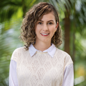 La imagen muestra a una persona en un entorno exterior con vegetación frondosa y tonos verdes desenfocados al fondo. La persona viste una camisa blanca de cuello clásico y, encima, un suéter sin mangas de tejido claro con un patrón calado. El cabello es rizado y de longitud media, peinado hacia un lado.