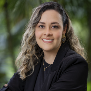 La imagen muestra a una persona en un entorno exterior con vegetación frondosa al fondo. La persona viste una chaqueta oscura sobre una prenda negra y lleva un collar delgado en tono dorado. El cabello es largo, con ondas marcadas y mechones claros. También se observan aretes grandes de diseño ornamental.