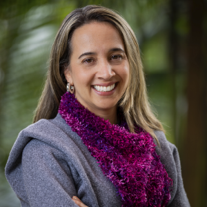 La imagen muestra a una persona en un entorno exterior con vegetación verde desenfocada al fondo. La persona viste una prenda de abrigo gris y una bufanda de textura esponjosa en tonos fucsia y morado brillante. También se observan aretes metálicos y parte del cabello peinado hacia un lado.