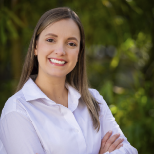 La imagen muestra a una persona de pie en un entorno exterior con vegetación verde desenfocada al fondo. La persona viste una camisa blanca de manga larga y mantiene los brazos cruzados. El cabello es lacio y de longitud media.