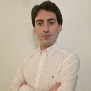 La imagen muestra a una persona de pie frente a una pared clara, vistiendo una camisa blanca con un patrón de líneas delgadas y los brazos cruzados. El cabello es de longitud media y el fondo es sencillo y uniforme.