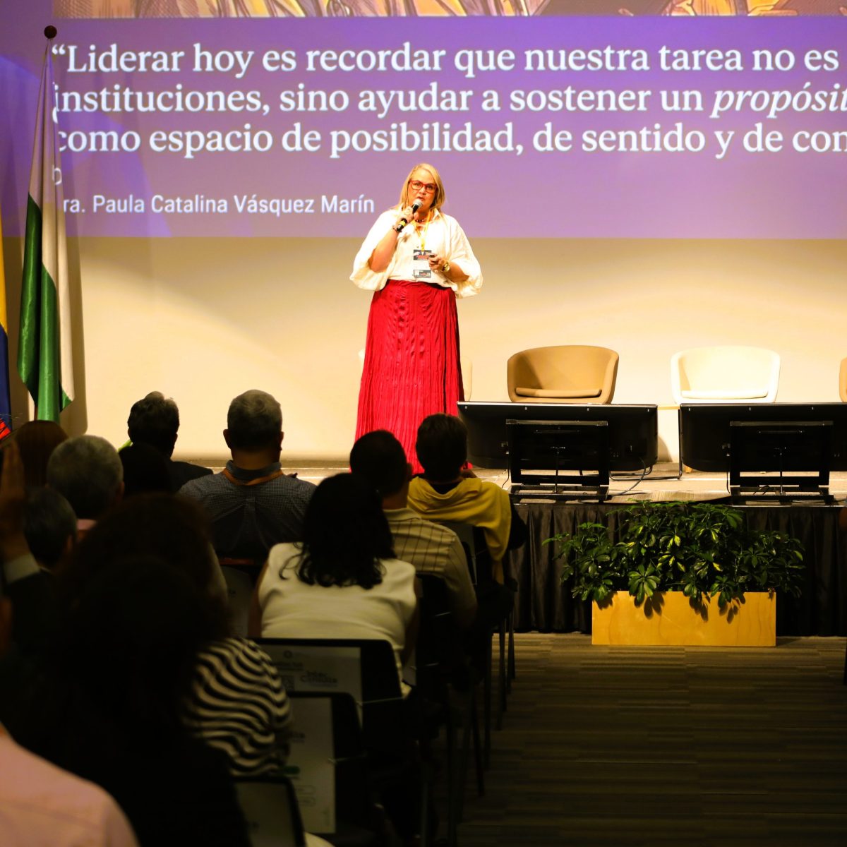 Persona dando una conferencia en un auditorio, de pie frente a una audiencia sentada. Detrás, una pantalla proyecta una frase sobre el liderazgo y el propósito institucional; en el escenario hay sillas y pantallas, y a un costado se observa una bandera.