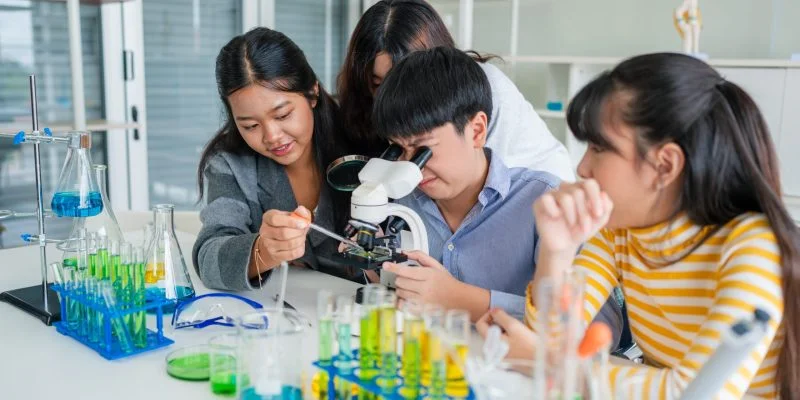 Grupo de estudiantes trabajando en un laboratorio escolar, observando muestras con un microscopio y manipulando tubos de ensayo con líquidos de colores.
