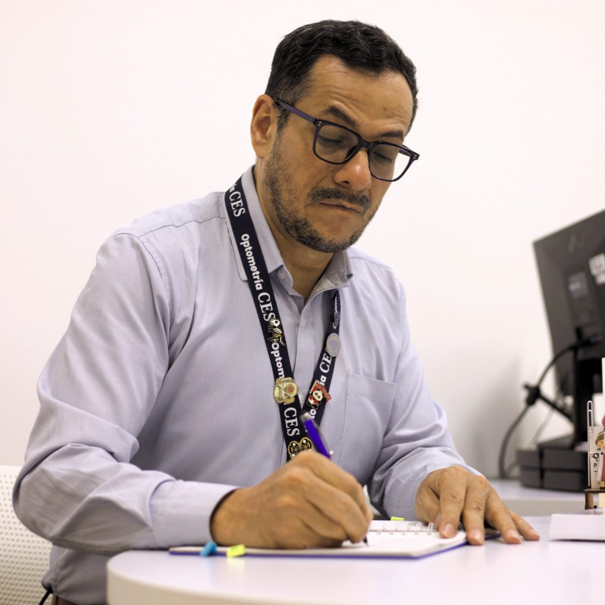 Persona sentada frente a un escritorio, escribiendo en un cuaderno con un bolígrafo. Lleva gafas y una credencial colgada al cuello; sobre la mesa se observan útiles de escritura y, al fondo, un equipo electrónico.