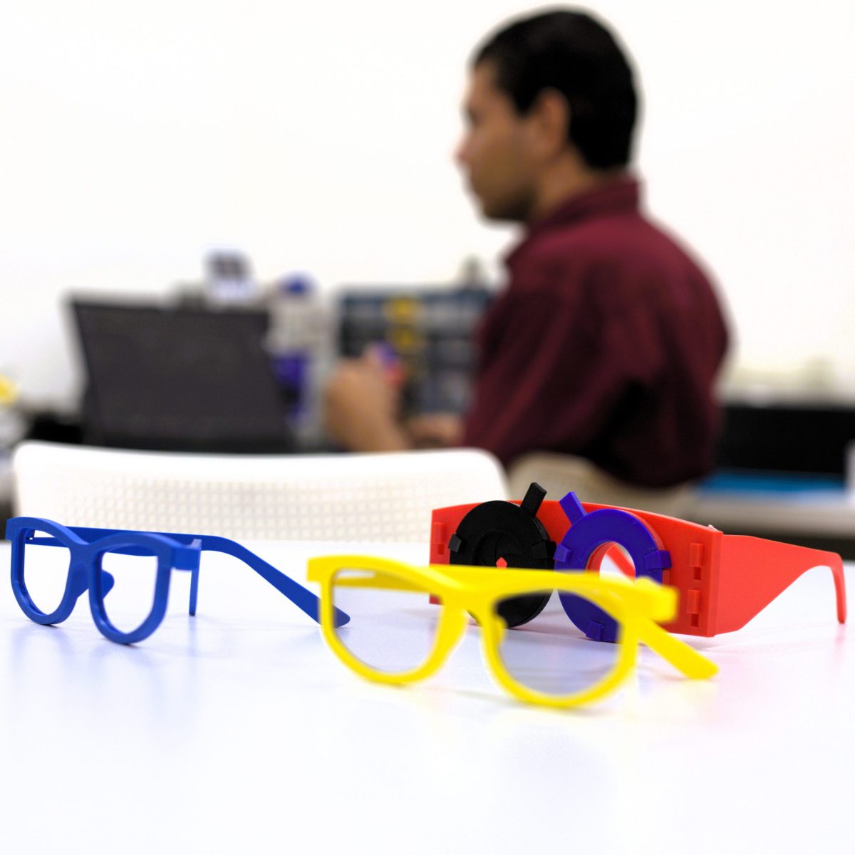 En primer plano, varias monturas de gafas de colores colocadas sobre una mesa blanca. Al fondo, una persona desenfocada sentada frente a un computador en un entorno interior de laboratorio.