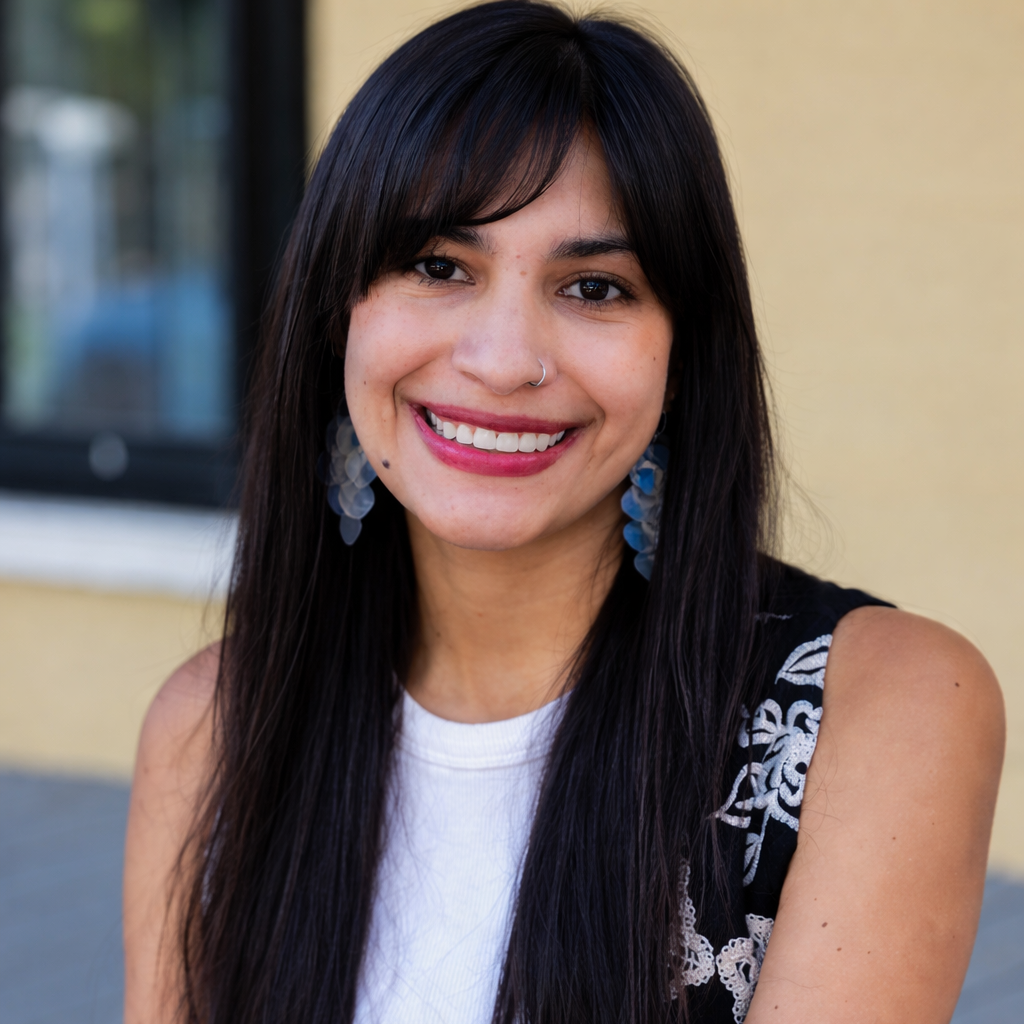 Retrato de una persona con cabello largo oscuro y flequillo, mirando a la cámara, con arete en la nariz, pendientes colgantes y blusa clara, sobre un fondo desenfocado.