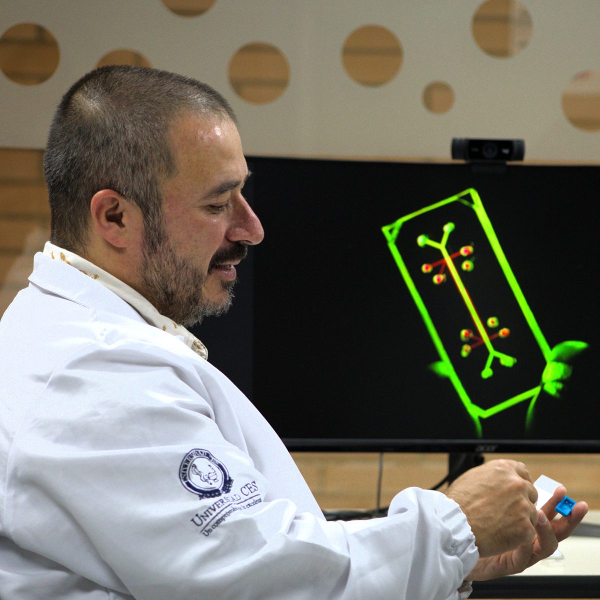 Persona con bata de laboratorio sostiene un pequeño dispositivo azul frente a una mesa, con una pantalla al fondo que muestra un esquema gráfico en color verde.