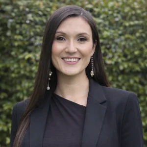 La imagen muestra a una persona de frente, con el cabello largo, lacio y oscuro, peinado hacia un lado. Viste un saco negro sobre una camiseta oscura y usa aretes largos con detalles colgantes. El fondo es exterior, con vegetación verde desenfocada y luz natural uniforme. Envía tus comentarios en BizChat