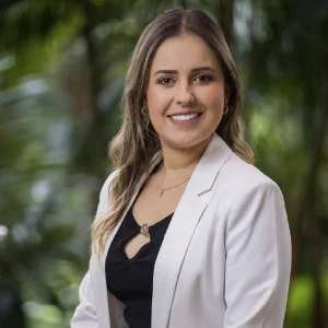La imagen muestra a una persona de frente en un entorno exterior con abundante vegetación verde desenfocada al fondo. Viste un blazer claro sobre una blusa oscura con escote en “V” y lleva un collar delicado. El cabello es largo, suelto y ligeramente ondulado. La iluminación es natural y uniforme.