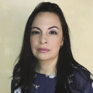 La imagen muestra a una persona de frente frente a un fondo liso de color claro. Tiene el cabello largo, oscuro y lacio, peinado hacia los lados. Viste una blusa de color azul oscuro con un diseño discreto en tonos claros. La iluminación es uniforme y el encuadre es de primer plano.