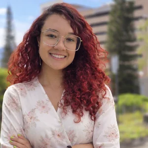 La imagen muestra a una persona de frente en un entorno exterior con fondo urbano y áreas verdes desenfocadas. Tiene el cabello rizado de tono rojizo y usa gafas de armazón claro. Viste una blusa clara con estampado floral y mantiene los brazos cruzados. La iluminación es natural y uniforme.