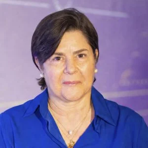 La imagen muestra a una persona de frente, en un entorno interior con fondo de color púrpura desenfocado. La persona viste una blusa azul de cuello abierto, usa aretes pequeños tipo perla y un collar delgado. El cabello es corto, oscuro y peinado hacia un lado. La iluminación es uniforme y el encuadre es de primer plano.
