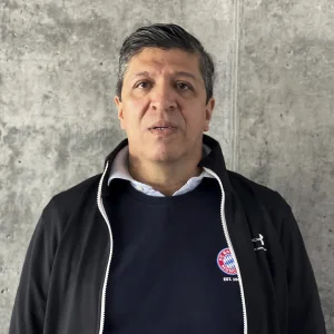 La imagen muestra a una persona de frente, en un entorno interior, frente a una pared de concreto gris con textura visible. Viste una chaqueta negra abierta sobre un suéter oscuro con un logotipo circular en el lado izquierdo del pecho y una camisa clara con cuello. La iluminación es uniforme y el encuadre es de medio cuerpo.
