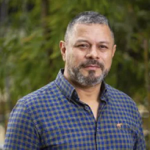 Persona con cabello corto y canoso, vistiendo camisa de cuadros azul y verde con cuello y botones, en un entorno exterior con vegetación desenfocada al fondo.