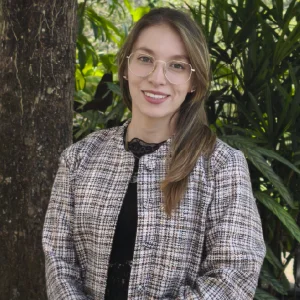La imagen muestra a una persona de pie en un entorno exterior, frente a abundante vegetación verde. Tiene el cabello largo y claro, recogido hacia un lado, y usa gafas de montura clara. Viste una chaqueta de tela con patrón claro sobre una prenda oscura. La iluminación es natural y el encuadre es de medio cuerpo.