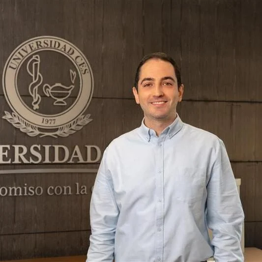 Persona de pie frente a un fondo institucional de madera con el escudo y el texto de una universidad; viste camisa clara de manga larga y se muestra centrada en la imagen.